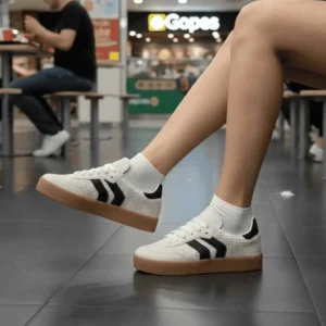 Tênis Feminino Casual Arrows na cor branca com listras laterais pretas e solado de borracha cor caramelo, sendo usado com calça social preta.