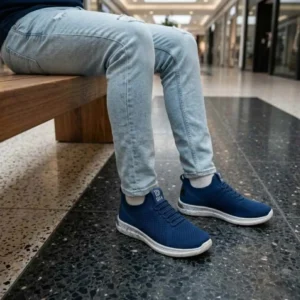 Tênis Masculino Casual Knit Conforto. Homem sentado em um banco de madeira usando tênis knit azul marinho e calça jeans clara em um ambiente interno iluminado.